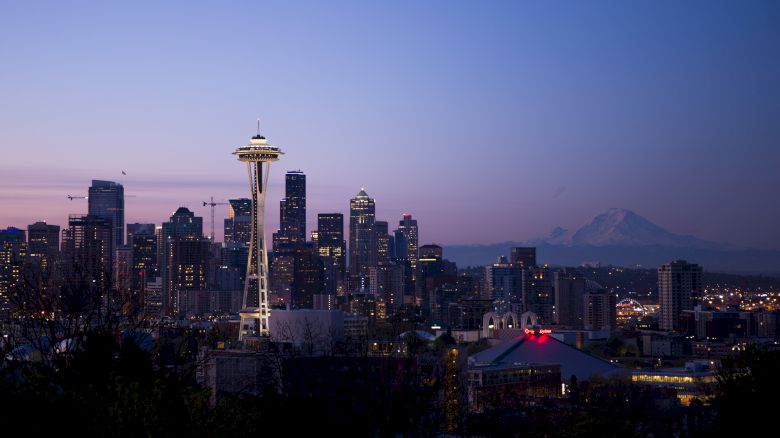 A city skyline at dusk featuring the Space Needle in the foreground and a mountain in the background, under a clear sky.