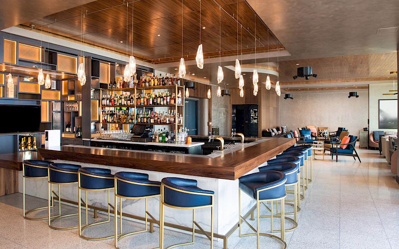 A modern bar with wooden accents, stylish lighting, ample seating, and a well-stocked selection of drinks.