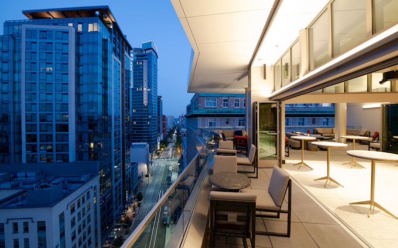 The image shows a modern cityscape at dusk with high-rise buildings and a rooftop terrace featuring outdoor seating and tables overlooking the city.