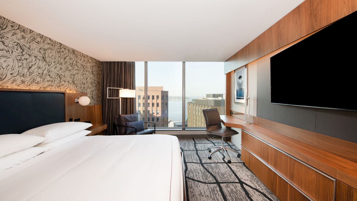 A modern hotel room with a large bed, desk, chair, and a wide-screen TV, featuring a window with a cityscape view in the background.