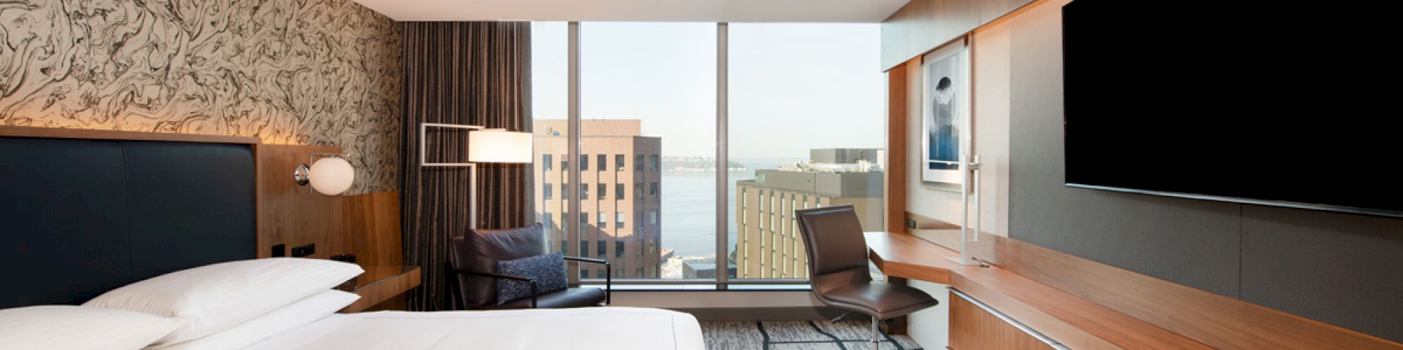 A modern hotel room with a large bed, desk, chair, and a wide-screen TV, featuring a window with a cityscape view in the background.