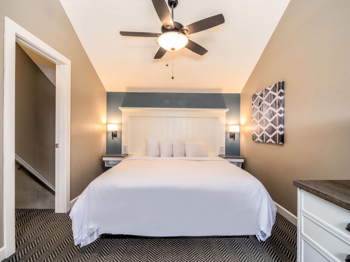 A neatly dressed bedroom with a ceiling fan, wall art, and a large bed with white linens and pillows in the center of the room.