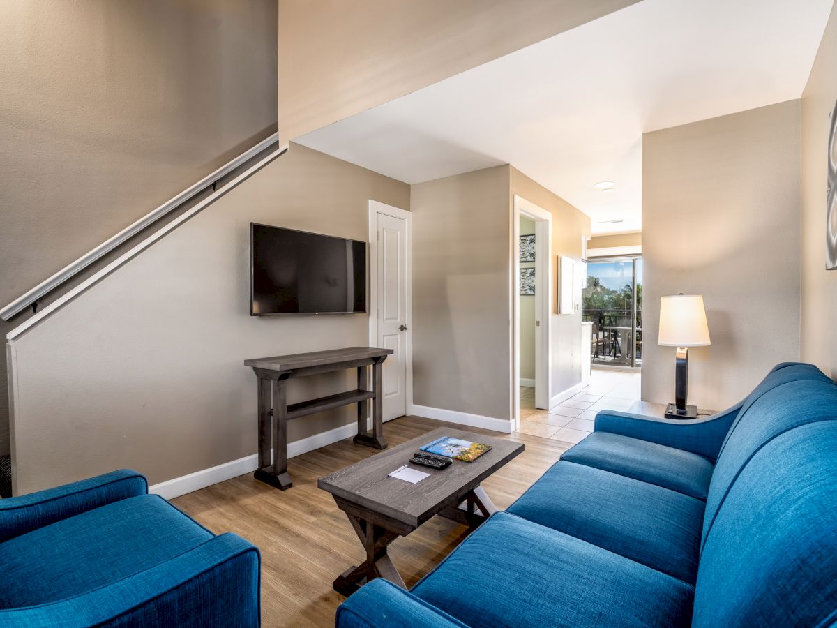 This image shows a cozy living room with blue seating, a TV on the wall, a wooden coffee table, a lamp, and a staircase leading upstairs.