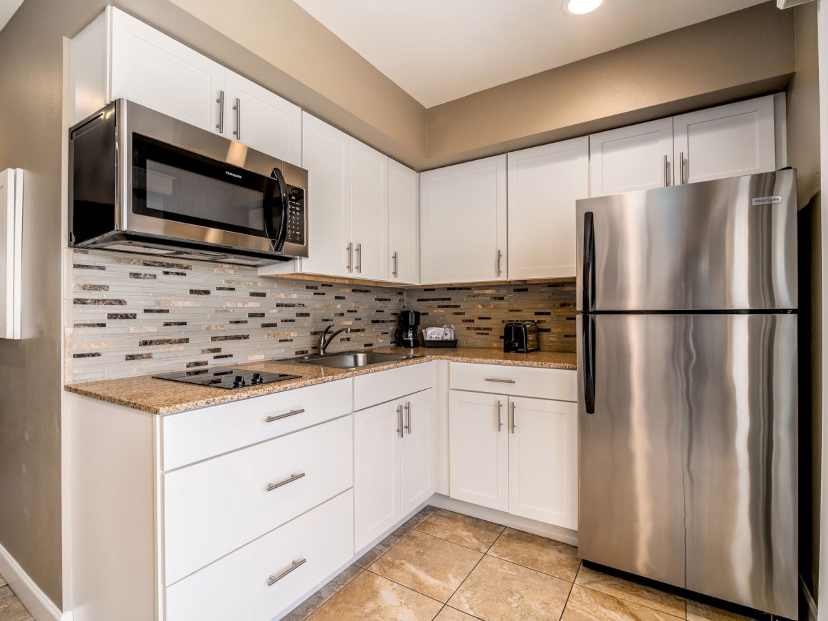 A modern kitchen with white cabinets, a stainless steel refrigerator, microwave, and a tiled backsplash. The counter has a toaster and a coffee maker.