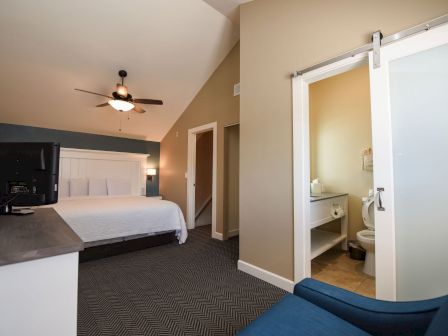 A modern hotel room features a bed, ceiling fan, TV, armchair, and an adjacent bathroom with a sliding door.