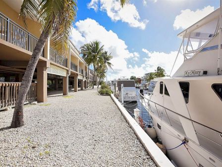 A marina with a docked yacht, palm trees, and buildings with balconies under a partly cloudy sky on a bright day.